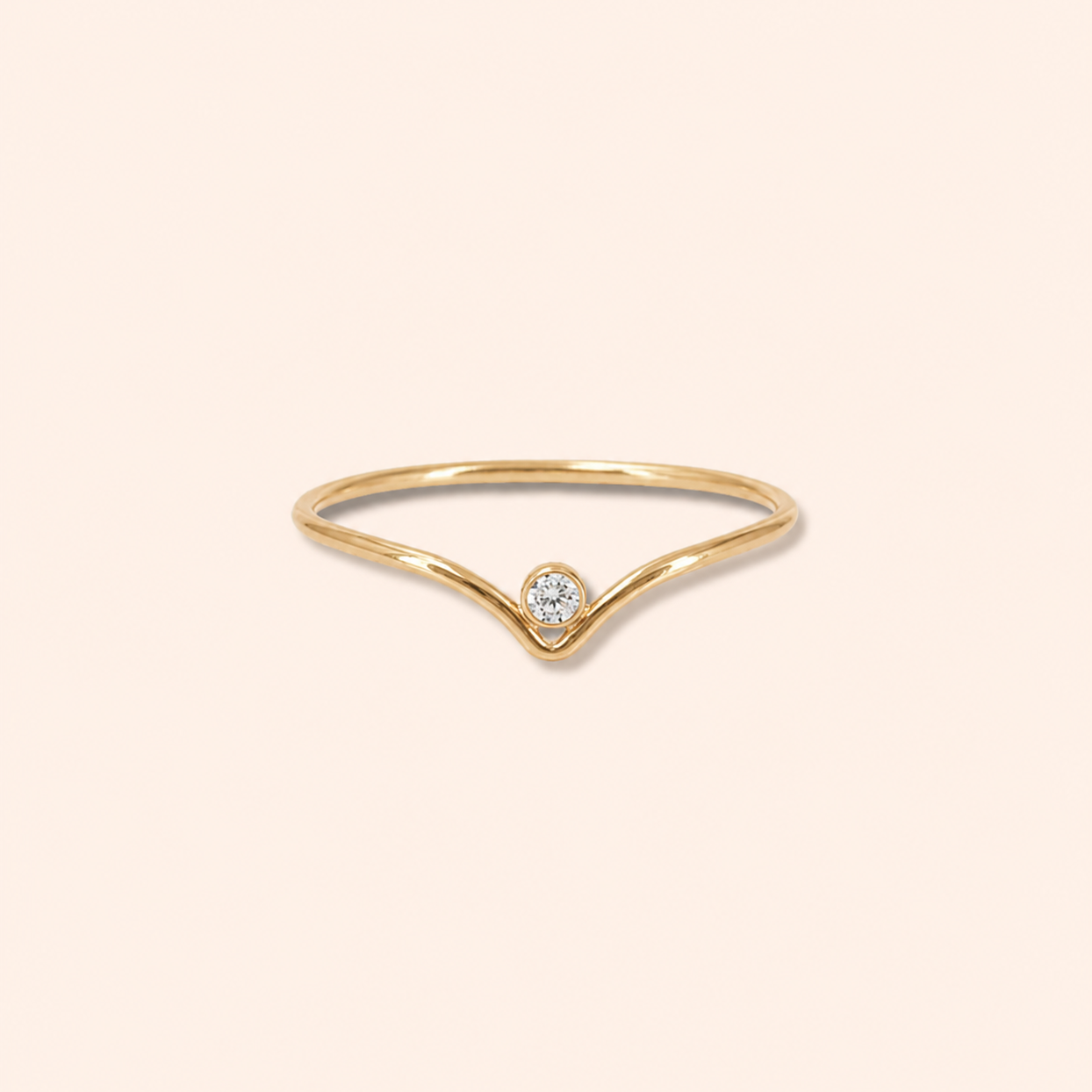 A delicate gold ring with a V-shaped band and a single round-cut clear gemstone at the center, set against a solid pale peach background.