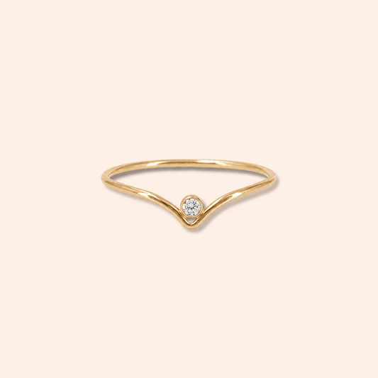 A delicate gold ring with a V-shaped band and a single round-cut clear gemstone at the center, set against a solid pale peach background.