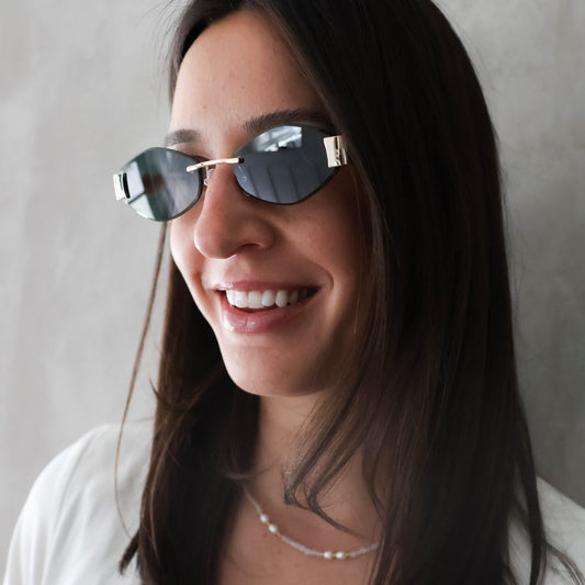 A woman with long dark hair wearing oval sunglasses with a silver metal frame and dark lenses, paired with a white top and a delicate pearl necklace, set against a neutral gray background.