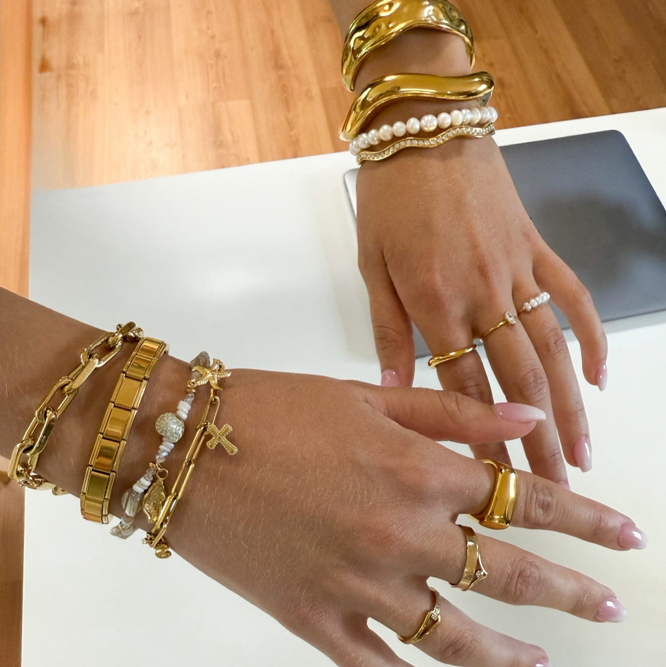 Two hands wearing gold bracelets and rings on a light background