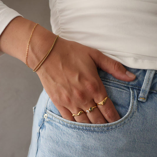A close-up lifestyle shot of a person wearing light blue denim jeans, a white top, and delicate gold jewelry, including two rings and a chain bracelet, resting their hand on their thigh.