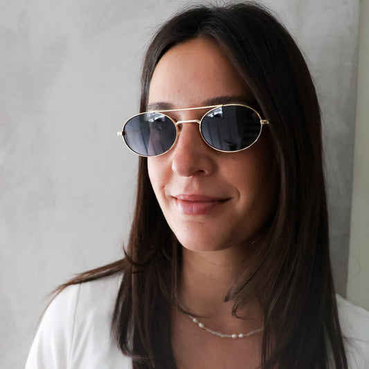A woman with long dark hair wearing gold-framed aviator sunglasses with dark lenses and a white top, photographed against a light gray textured wall.