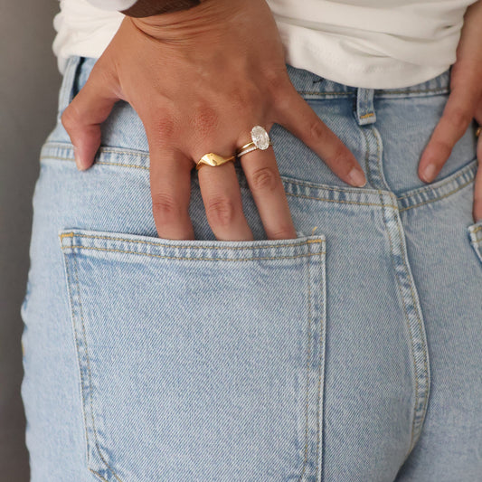 Close-up of a person wearing light blue jeans, a white long-sleeve top, and a watch, with two rings on their fingers resting in the back pocket.