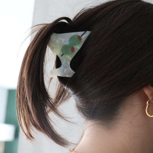 A woman shown from the back wearing a large, stylish hair claw clip with a green and white botanical pattern, holding her dark brown hair in a ponytail.