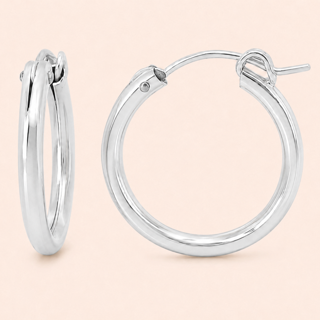A pair of polished silver hoop earrings shown on a solid pale peach background, with one earring standing upright and the other lying flat to display the front and latch mechanism.
