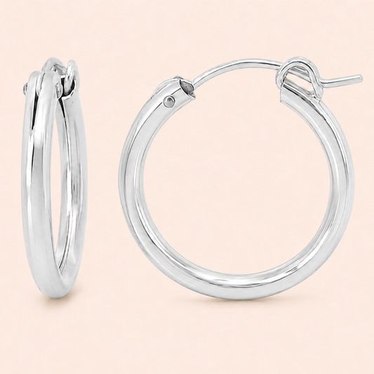 A pair of polished silver hoop earrings shown on a solid pale peach background, with one earring standing upright and the other lying flat to display the front and latch mechanism.