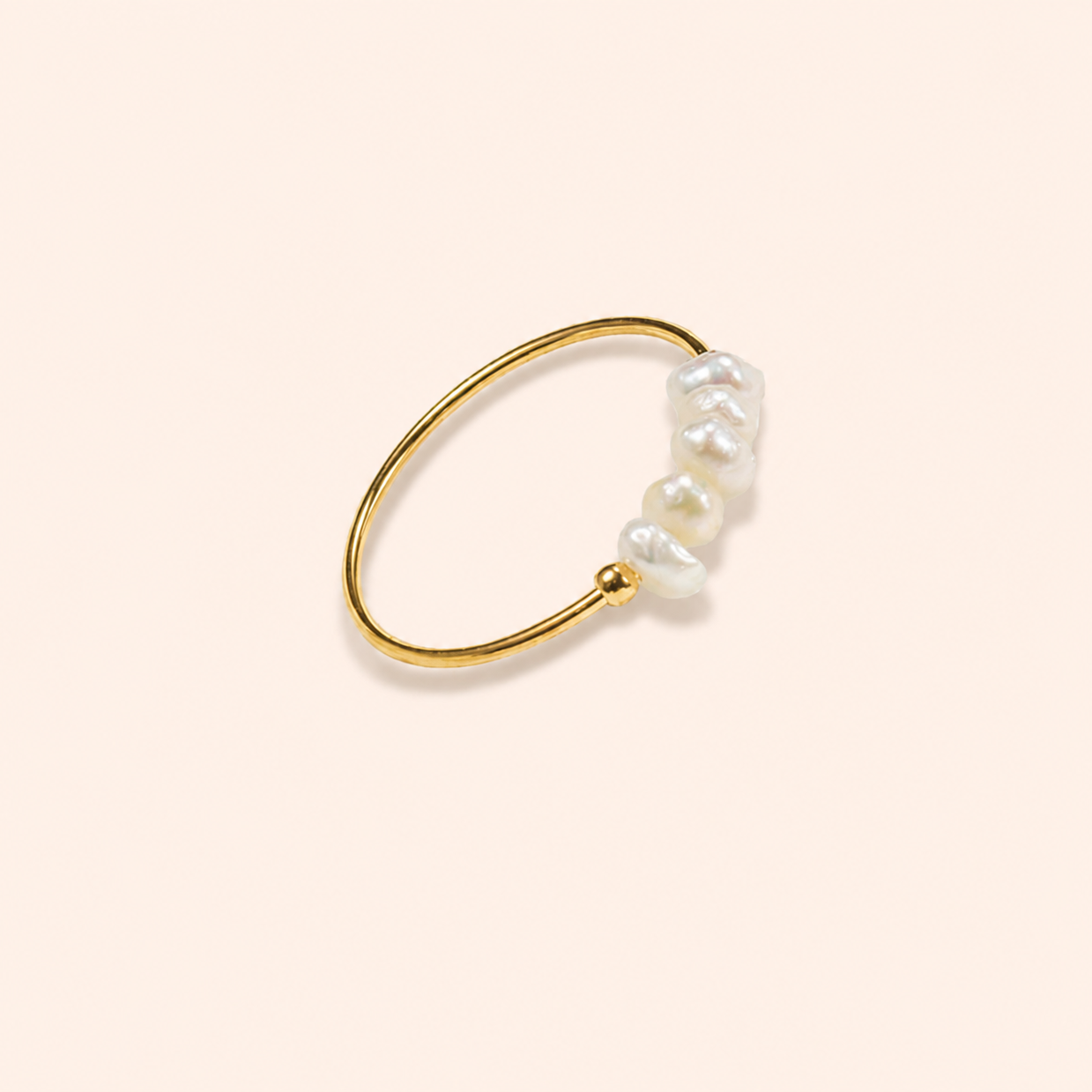 A delicate gold ring featuring a cluster of irregular white baroque pearls on one end, displayed on a solid pale peach background.