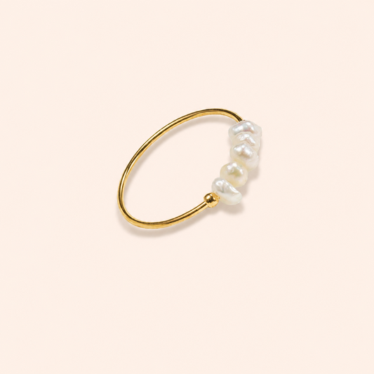 A delicate gold ring featuring a cluster of irregular white baroque pearls on one end, displayed on a solid pale peach background.