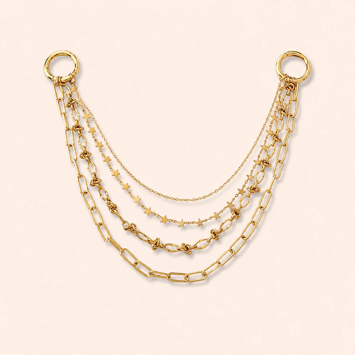 A gold-toned layered chain accessory with two large ring connectors at the top, featuring multiple draped chains including paperclip links, star motifs, and delicate cable chains on a pale peach background.
