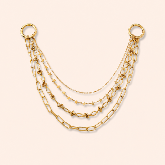 A gold-toned layered chain accessory with two large ring connectors at the top, featuring multiple draped chains including paperclip links, star motifs, and delicate cable chains on a pale peach background.