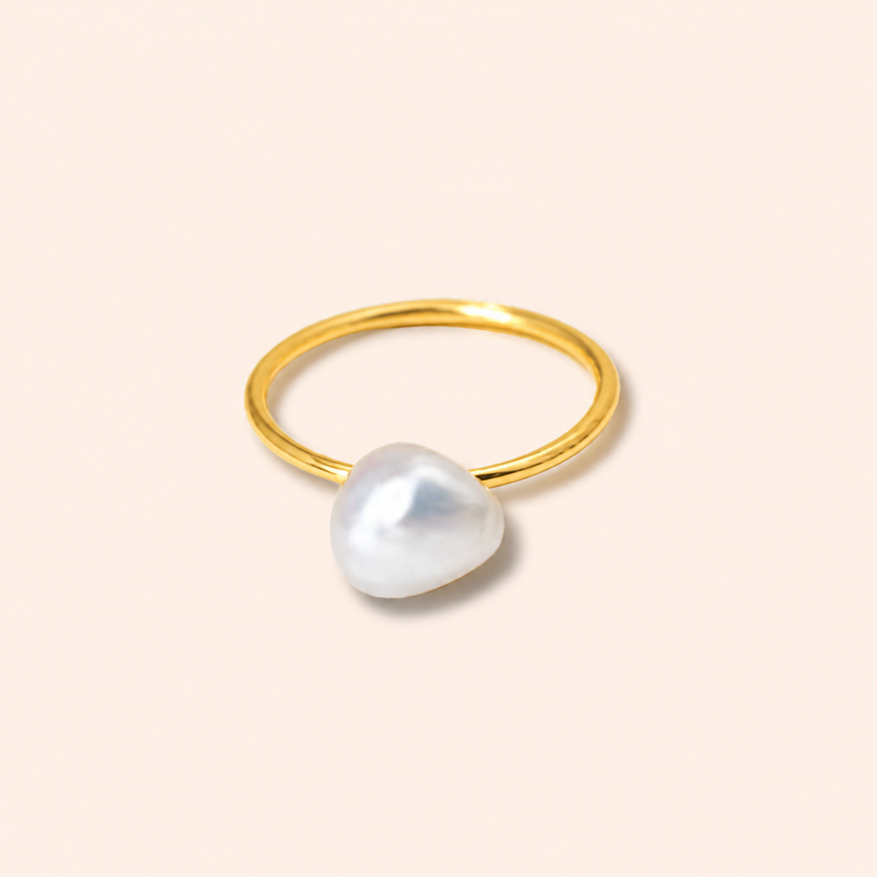 A minimalist gold ring featuring a large, irregular white baroque pearl as the centerpiece, set against a solid pale peach background.