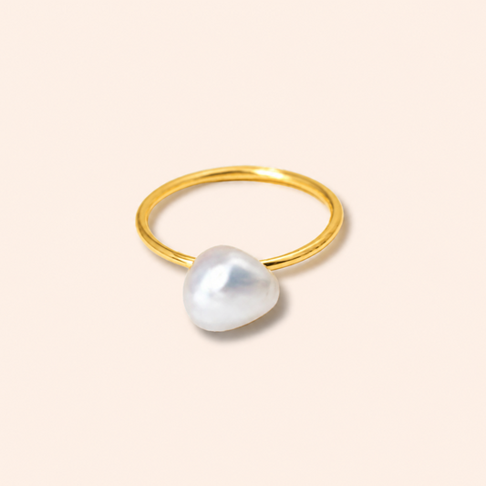 A minimalist gold ring featuring a large, irregular white baroque pearl as the centerpiece, set against a solid pale peach background.