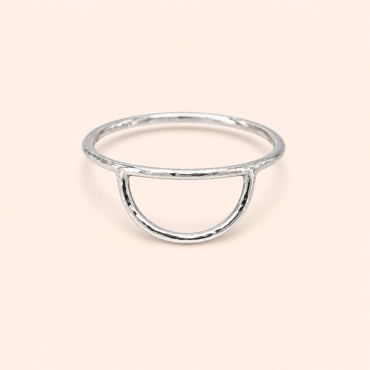 A minimalist silver ring with a hammered texture, featuring a semi-circular open arch design, set against a solid pale peach background.