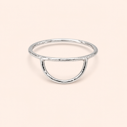 A minimalist silver ring with a hammered texture, featuring a semi-circular open arch design, set against a solid pale peach background.