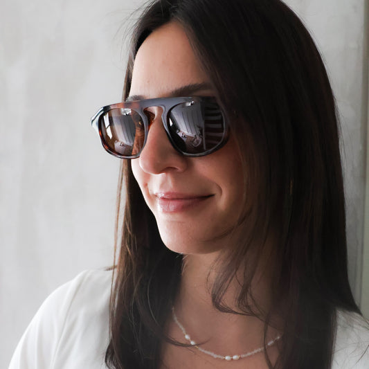 A woman with long dark hair wearing dark-framed sunglasses with brown tinted lenses and a white top, shown in a close-up portrait against a light gray wall.