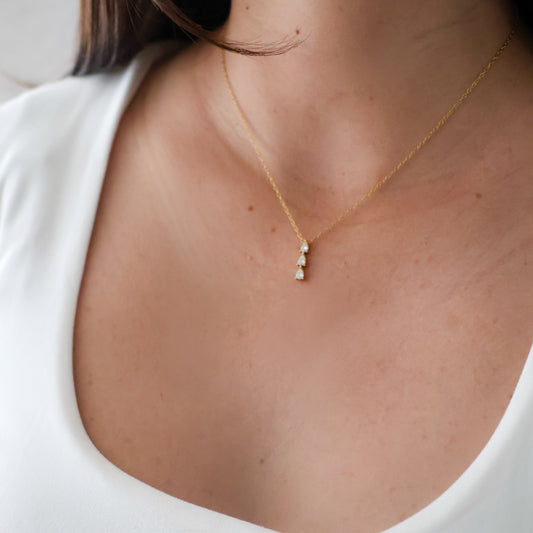 A close-up of a model wearing a delicate gold chain necklace with a small vertical cluster of white pearls at the center of the chest, paired with a white scoop-neck top.