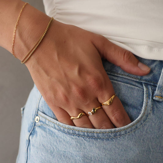 A close-up lifestyle shot of a person wearing light blue denim jeans, a white top, and delicate gold jewelry, including two rings and a chain bracelet, resting their hand on their thigh.