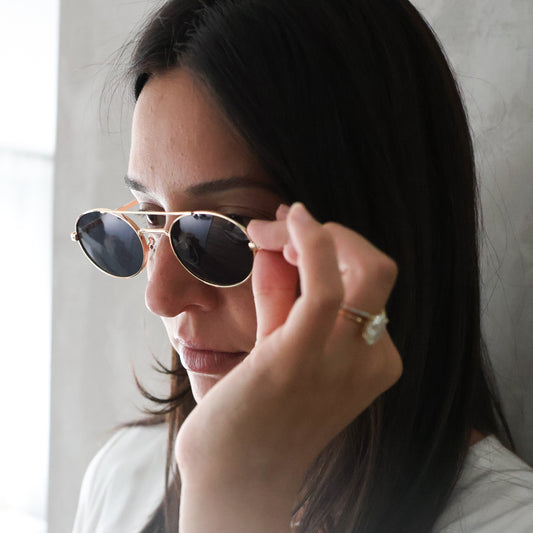 A woman with long dark hair wearing gold-framed aviator sunglasses with dark lenses, a white long-sleeve top, and a smartwatch, adjusting the bridge of her sunglasses with her right hand.