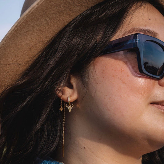 Close-up of a person wearing sunglasses, a hat, and a denim vest with visible jewelry.