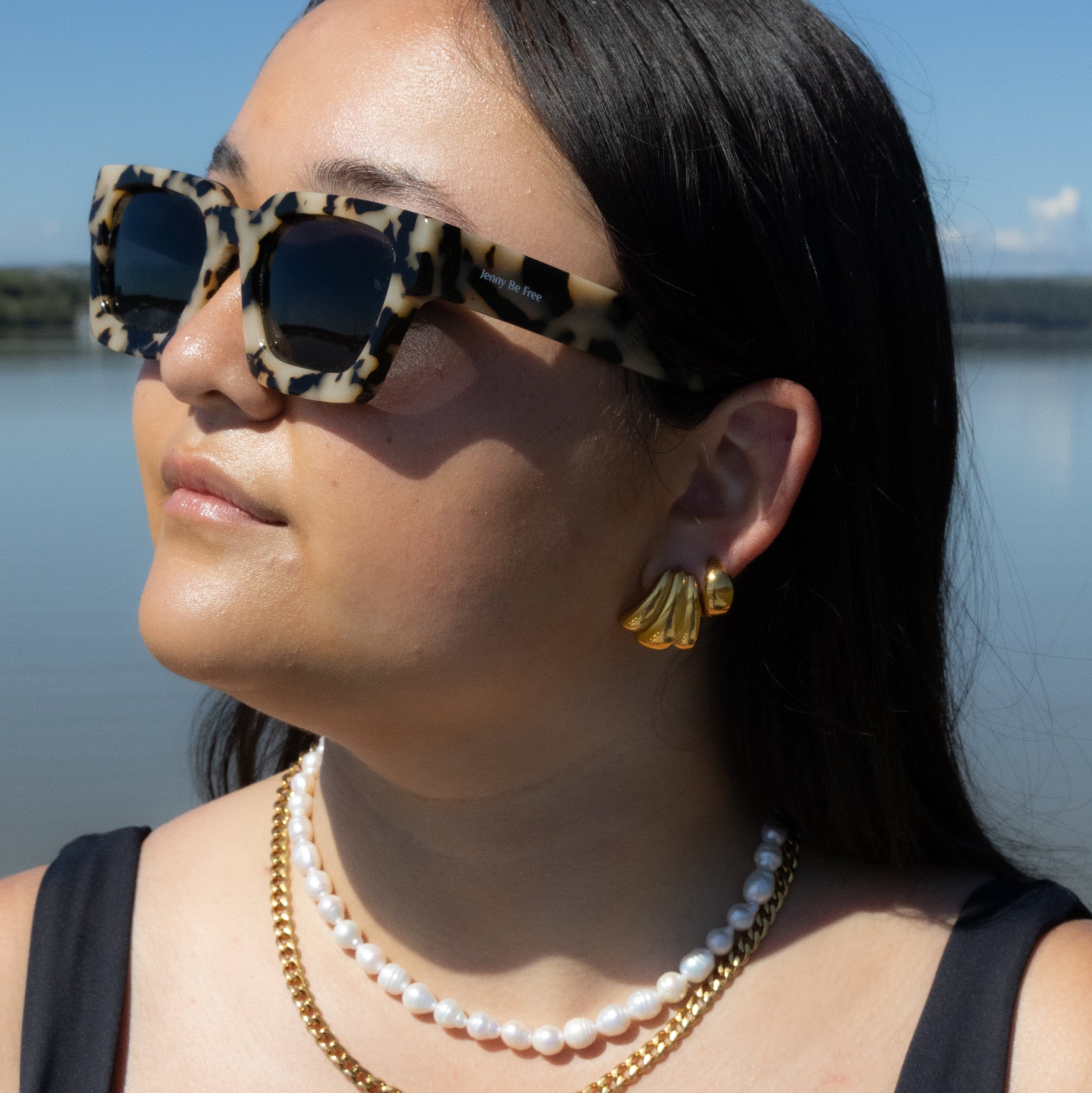 Woman wearing leopard print sunglasses and gold jewelry by a body of water.