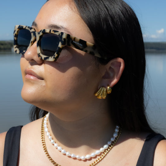 Woman wearing leopard print sunglasses and gold jewelry by a body of water.