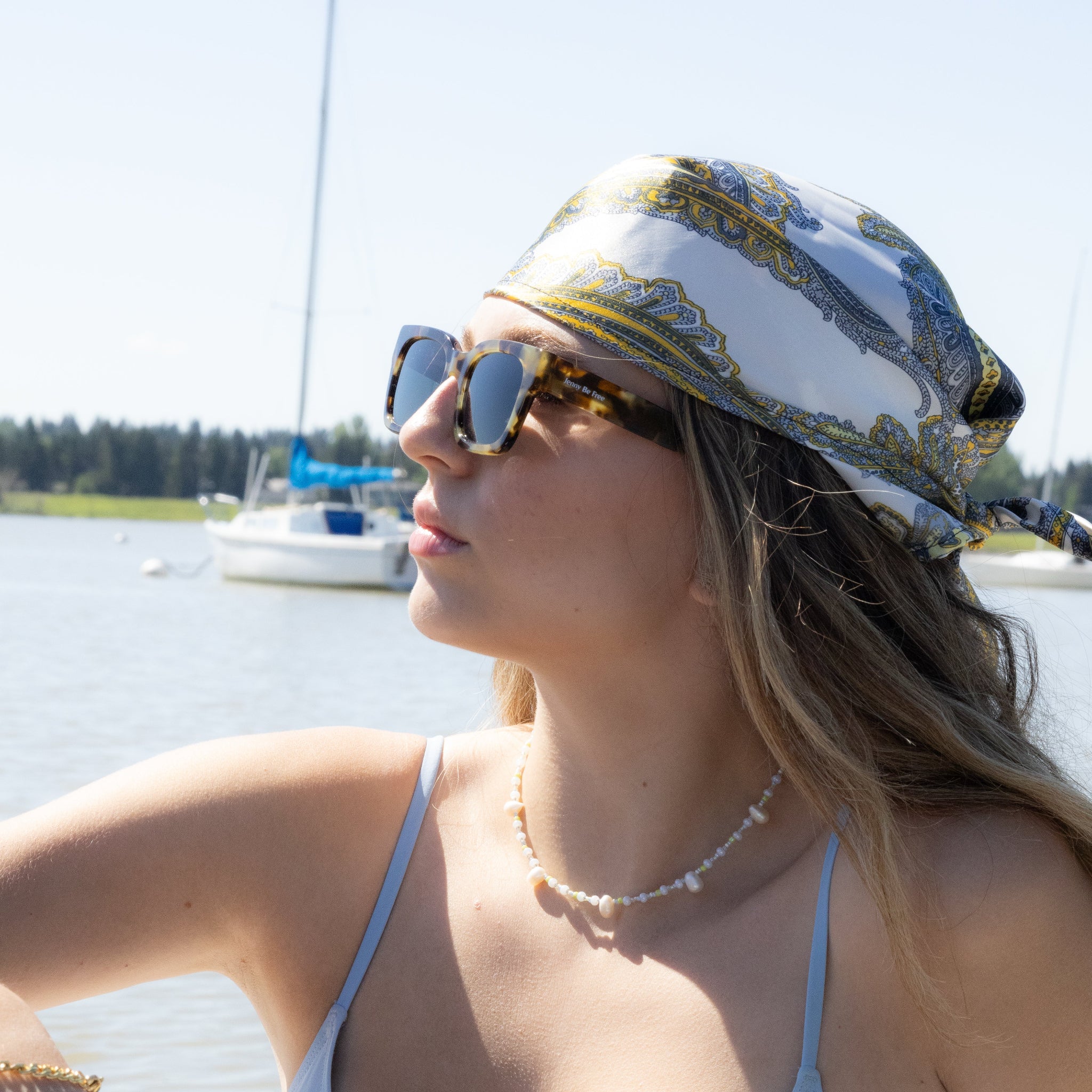 Woman wearing a white bikini top and patterned hat by a lake with boats in the background