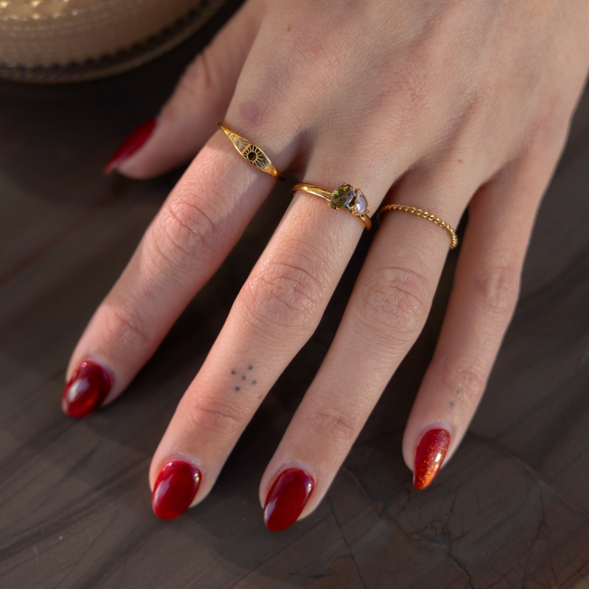 Hand with red nail polish wearing gold rings on a wooden surface with a blurred cup of coffee in the background.