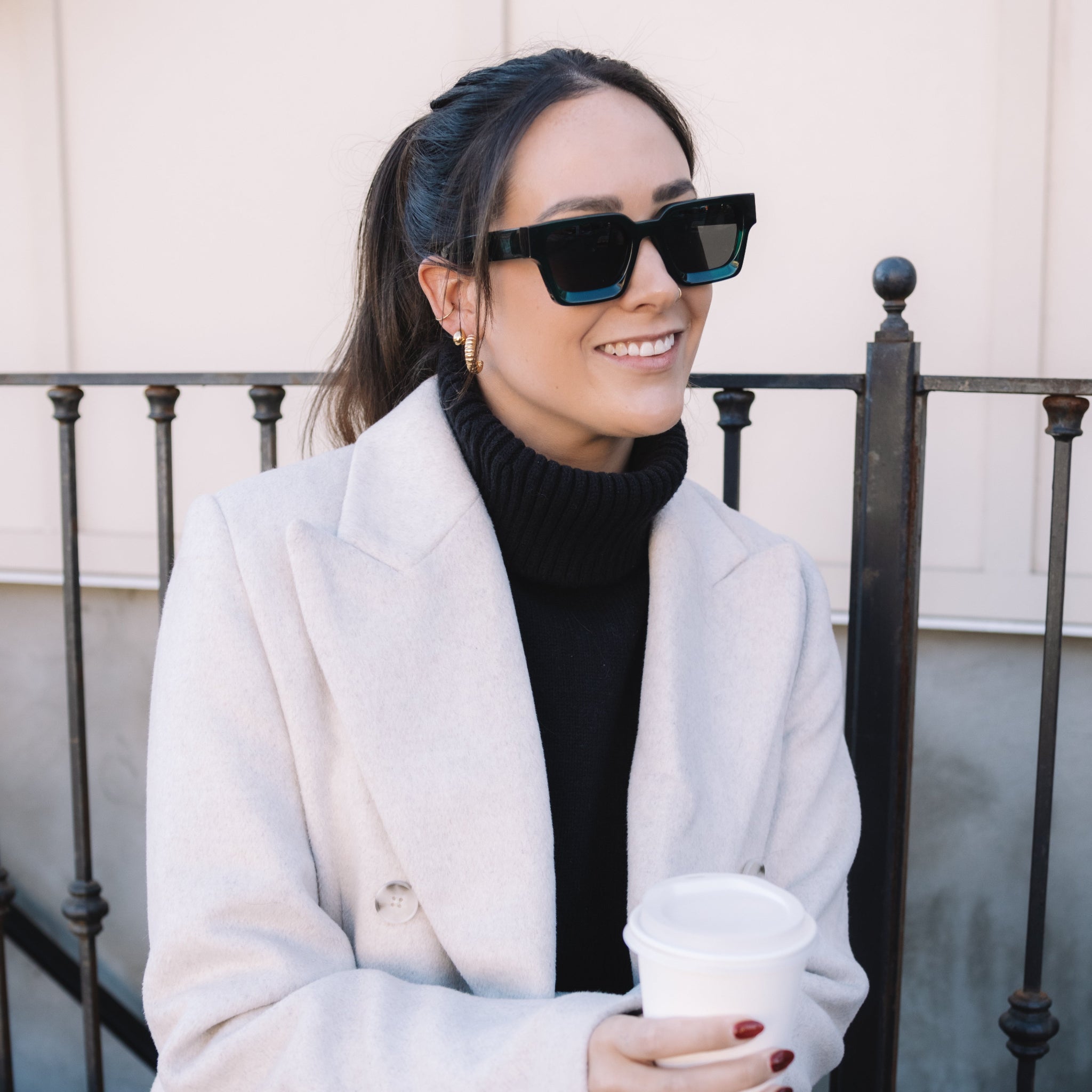 Woman wearing a white coat and sunglasses, sitting on a bench.
