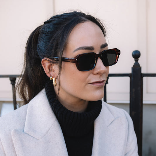 Woman wearing sunglasses and a light gray coat with a black background