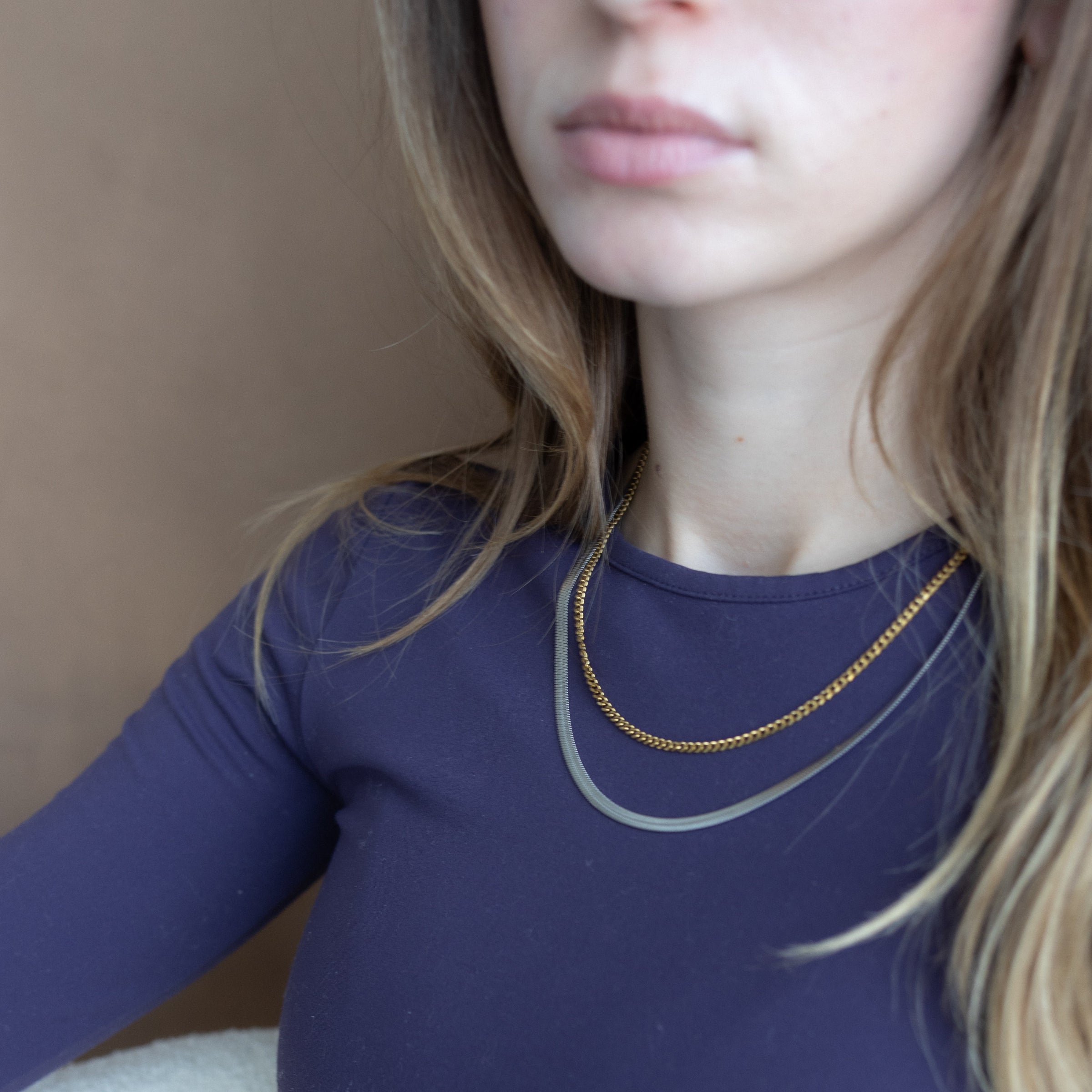 A close-up of a woman wearing a navy blue long-sleeve top and layered gold necklaces, with her face partially cropped at the top against a neutral background.