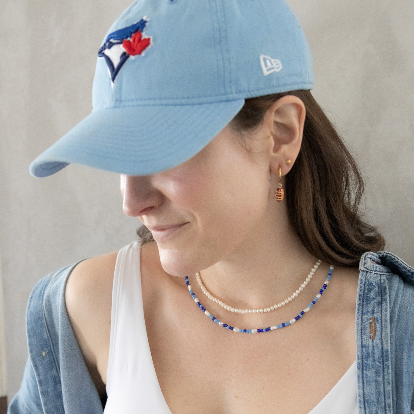 Woman wearing a blue baseball cap with a logo, white tank top, and denim jacket against a neutral background.