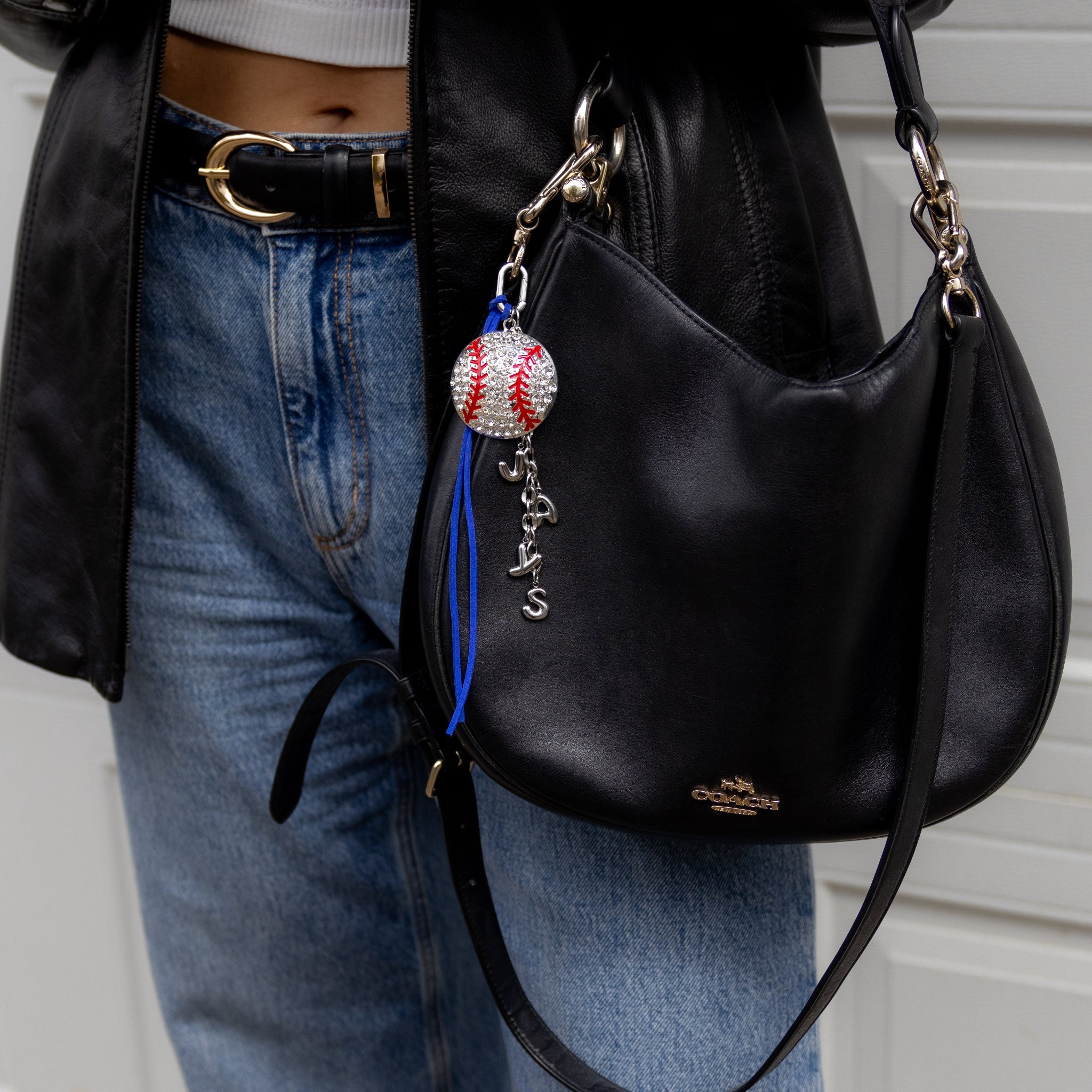 Person wearing a black leather jacket, white crop top, and blue jeans, holding a black handbag with a baseball charm.