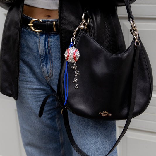 Person wearing a black leather jacket, white crop top, and blue jeans, holding a black handbag with a baseball charm.