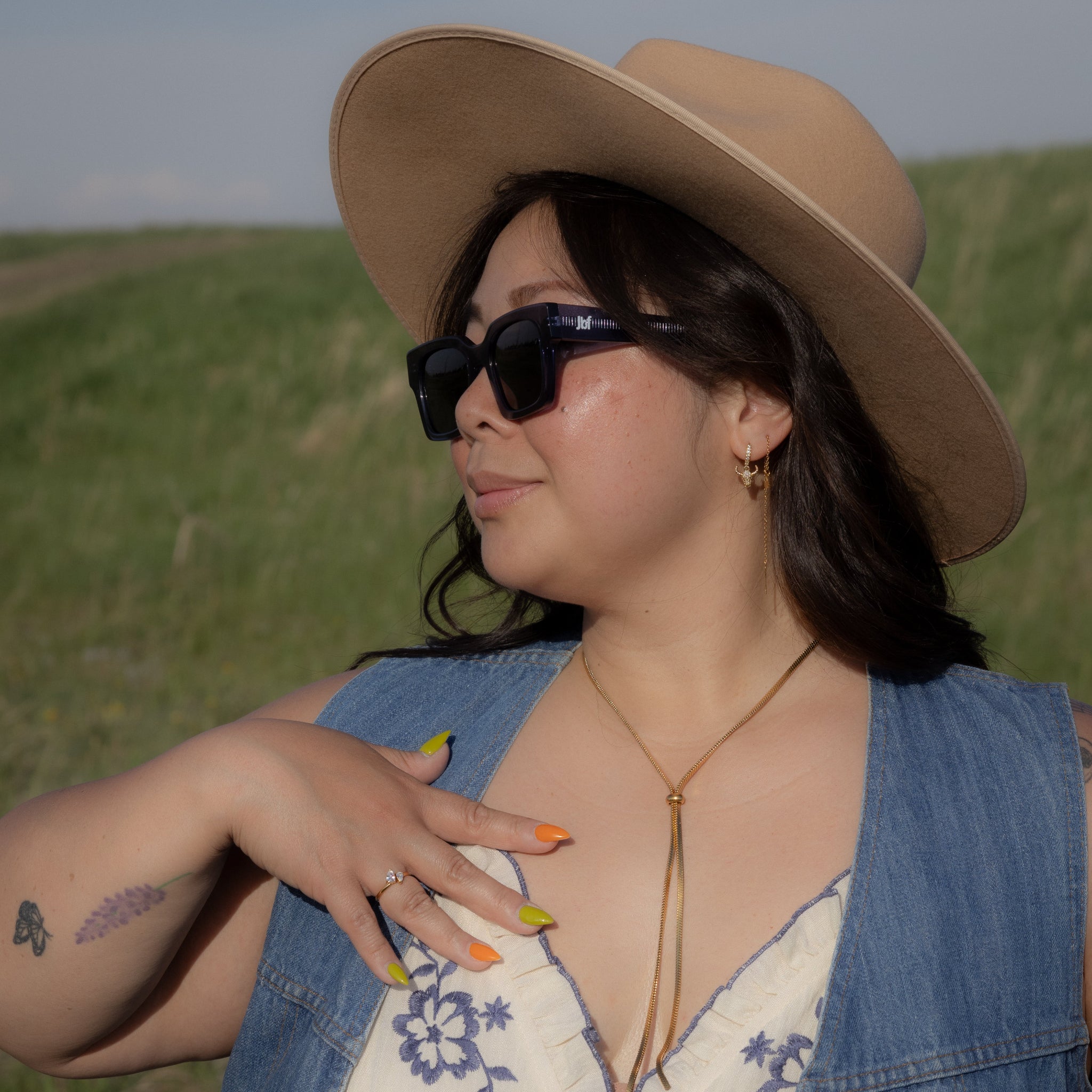 Woman wearing a beige hat, sunglasses, and a denim vest over a white dress with blue floral embroidery, standing in a grassy field.