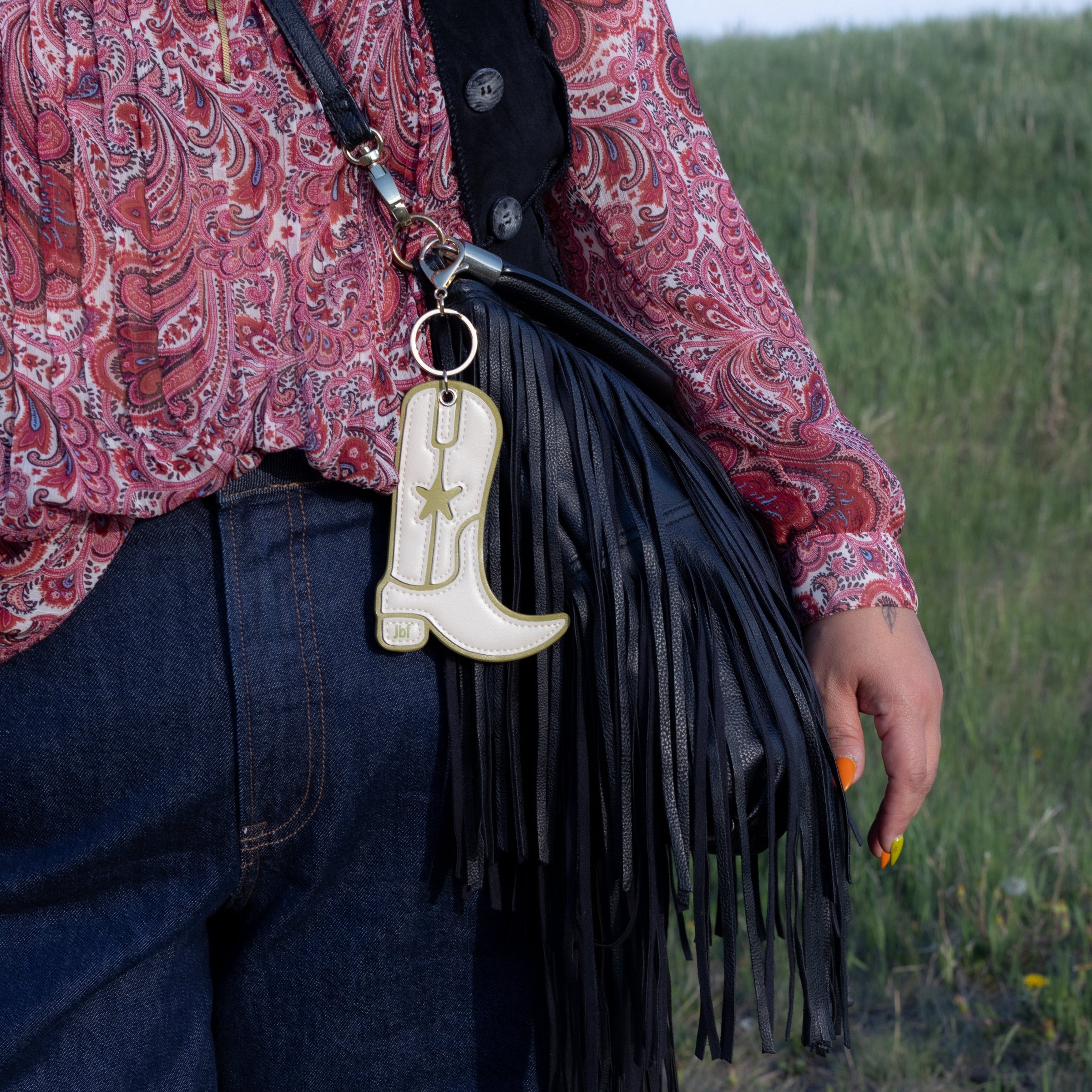 A close-up lifestyle shot of a person wearing a red paisley shirt and dark blue jeans, carrying a black leather fringe crossbody bag with a cream-colored faux cowhide boot-shaped keychain.