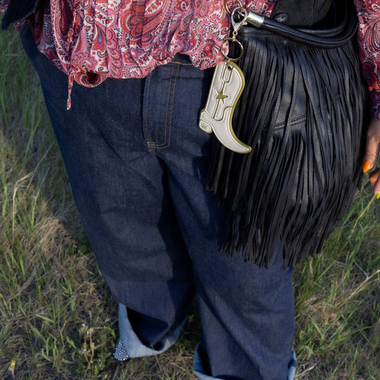 A person outdoors wearing dark blue jeans with rolled cuffs, a red paisley blouse, and black pointed boots, carrying a black fringe shoulder bag with a white cowboy boot-shaped charm.