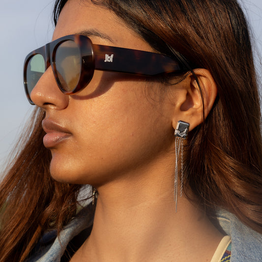 Woman wearing sunglasses and a denim jacket with a colorful top, against a light blue background