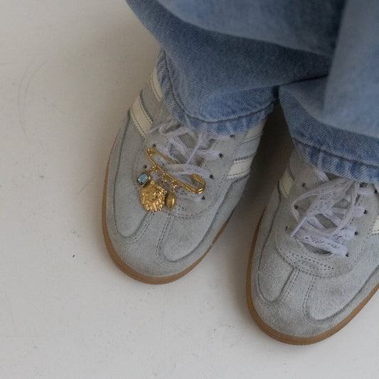 Close-up of a person's feet wearing light gray suede sneakers with light blue jeans, featuring a gold charm with a small blue gem on the laces of the left shoe.