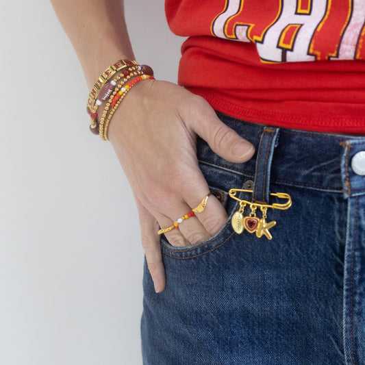 A close-up lifestyle shot of a person wearing a red graphic t-shirt and blue jeans, featuring a gold safety-pin style charm bracelet with a star and heart hanging from the front pocket.