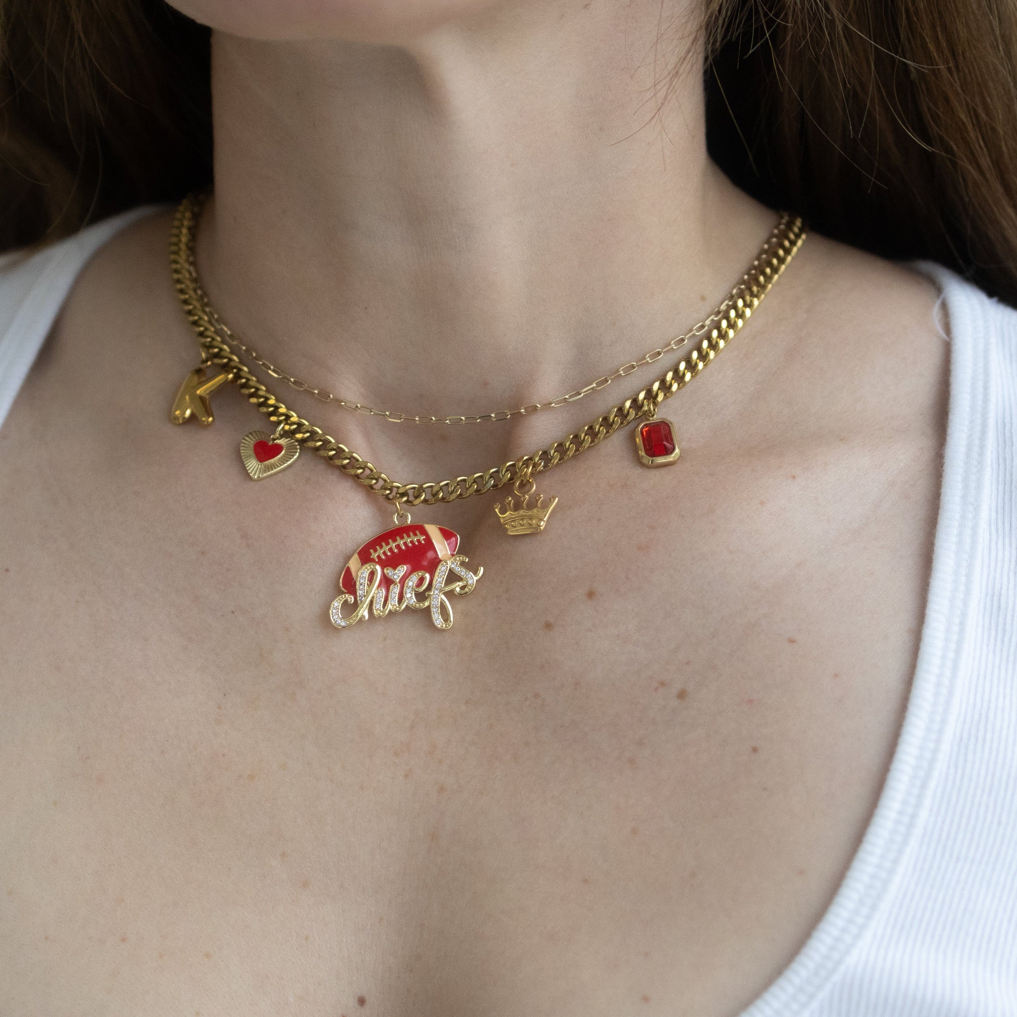 A close-up of a model wearing a layered gold necklace with red and gold charms, including a crown, small hearts, and a script charm spelling 'girl'.