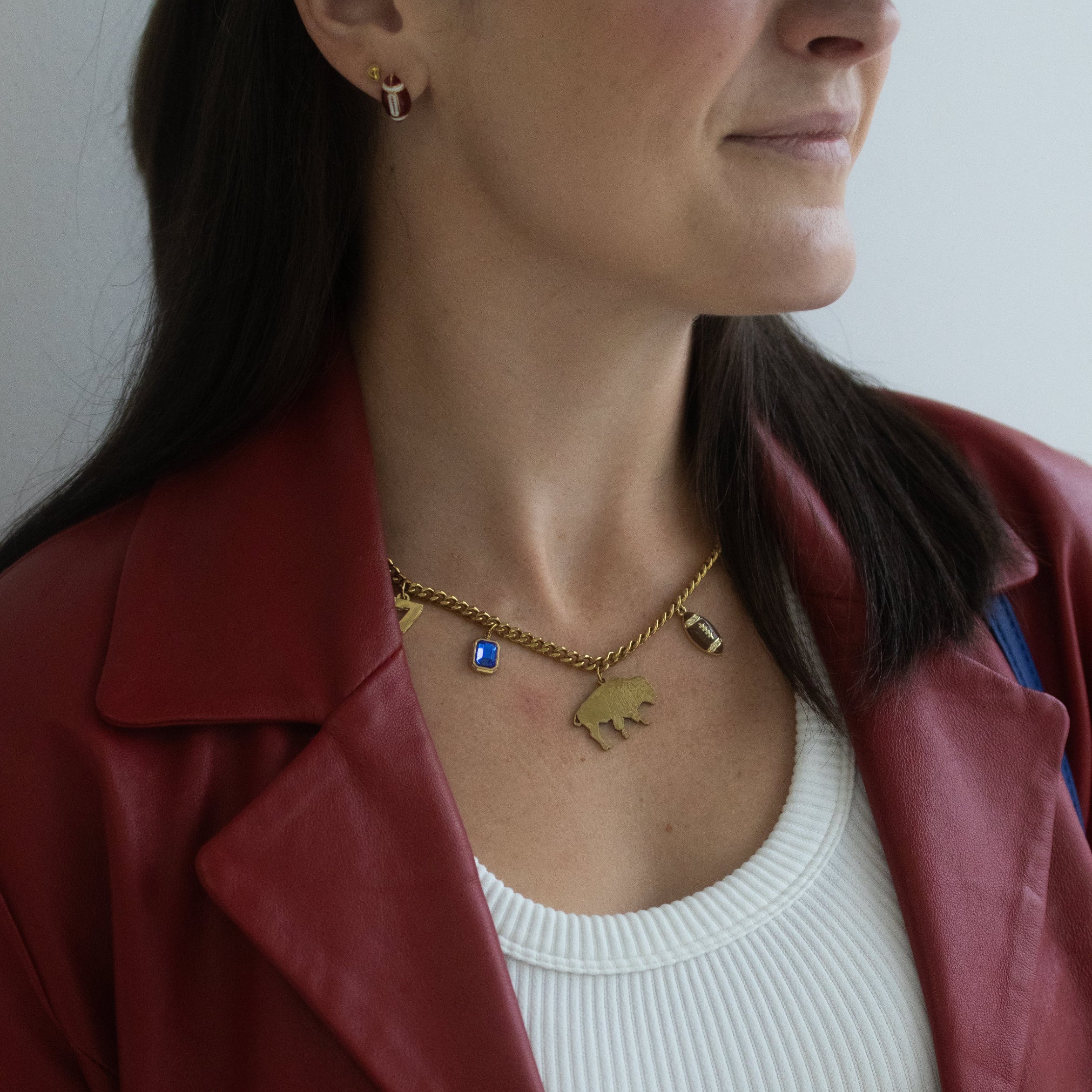 A close-up of a woman wearing a gold safety-pin style brooch with small heart charms, styled over a white ribbed top and a burgundy jacket.
