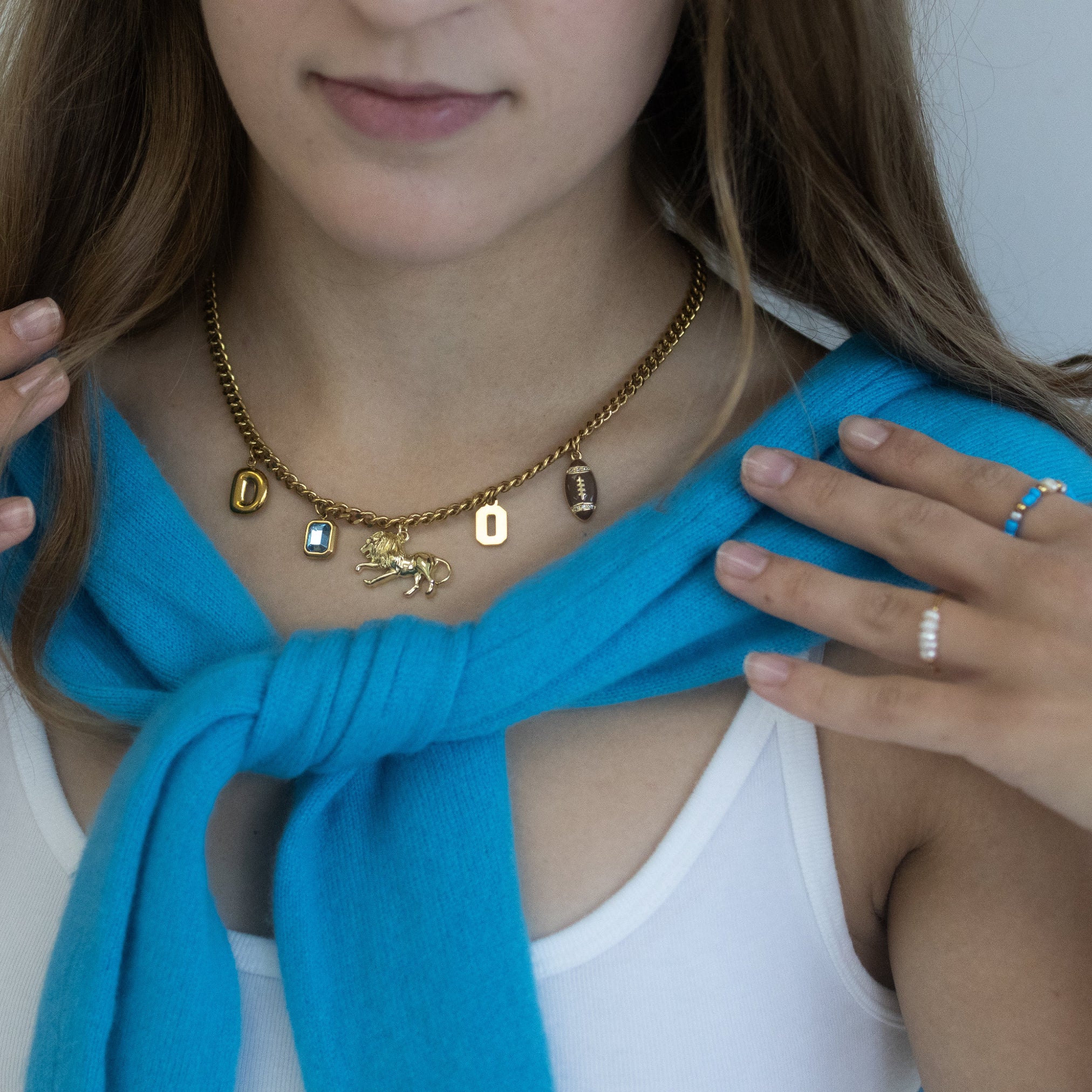 A close-up lifestyle shot of a woman wearing a gold letter charm necklace with a small animal charm, styled with a bright blue knit scarf and gold rings.