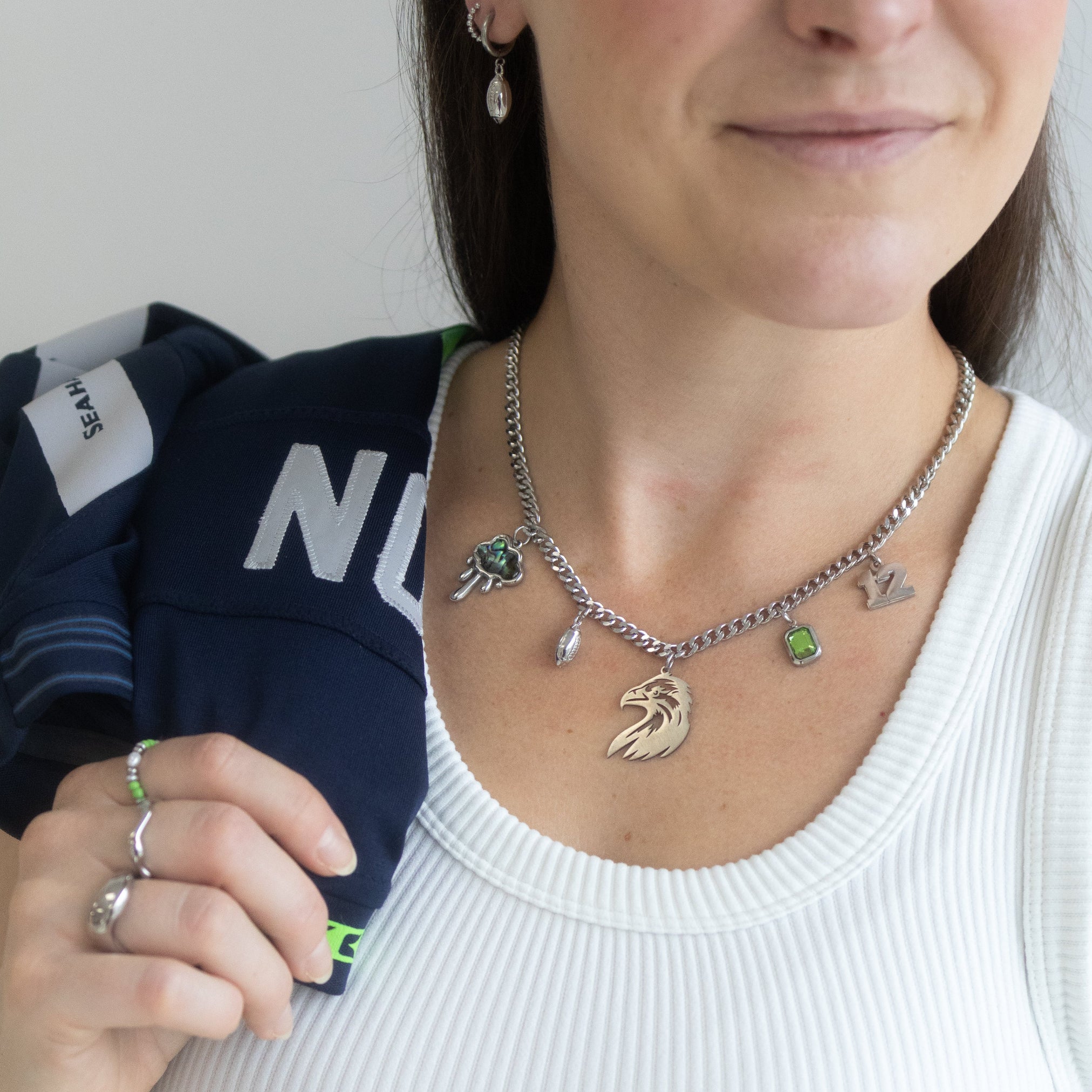 A close-up of a woman wearing a silver chain charm necklace with a horse head pendant and small letter charms, paired with a white ribbed tank top and holding a navy blue jacket.