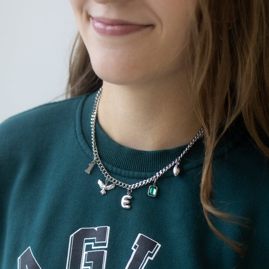 A close-up of a person wearing a silver-toned charm necklace with Eagles-themed charms, paired with a dark green Philadelphia Eagles sweatshirt.