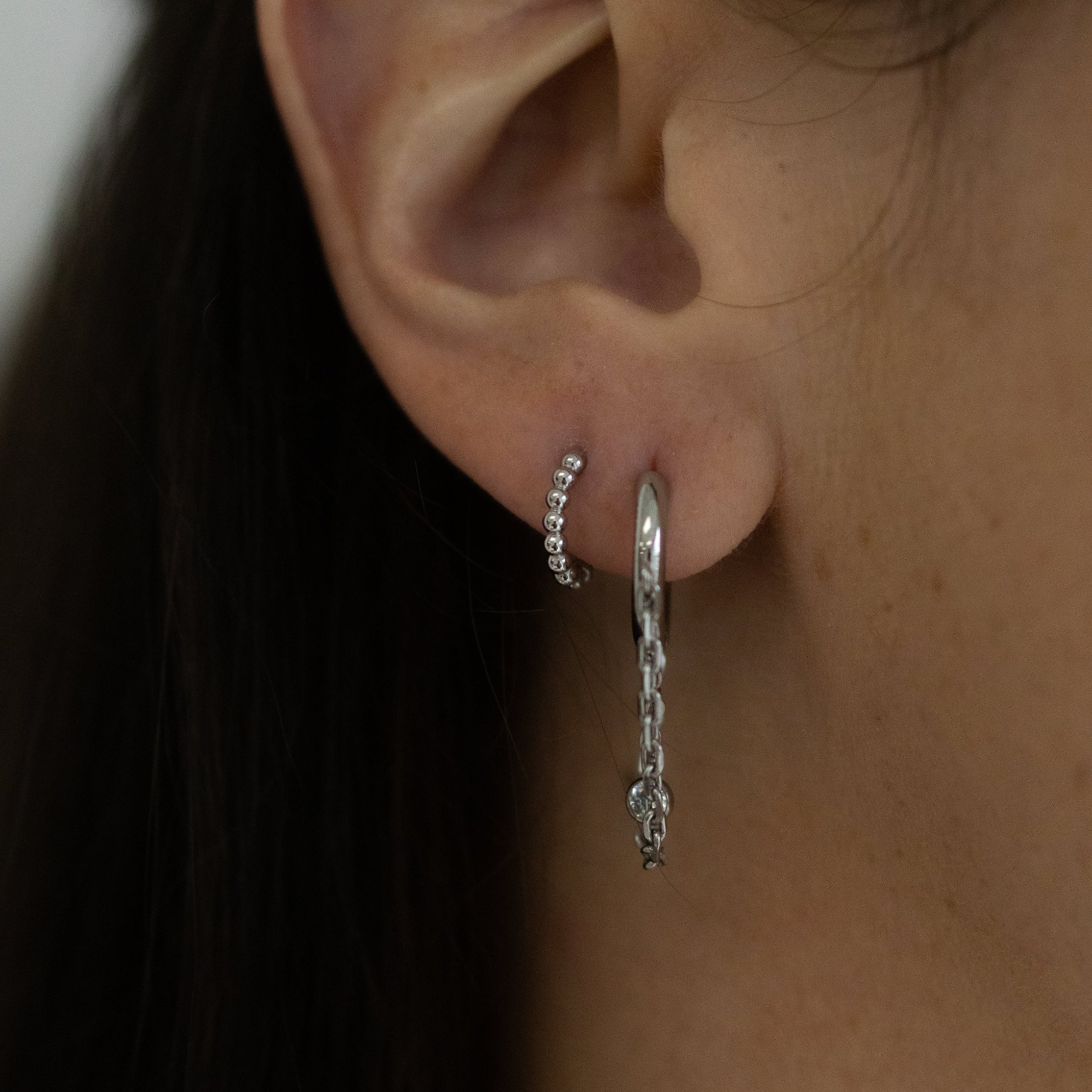 A close-up of a person's ear wearing two silver hoop earrings, one of which has a dangling chain with a small charm, against dark hair and skin.