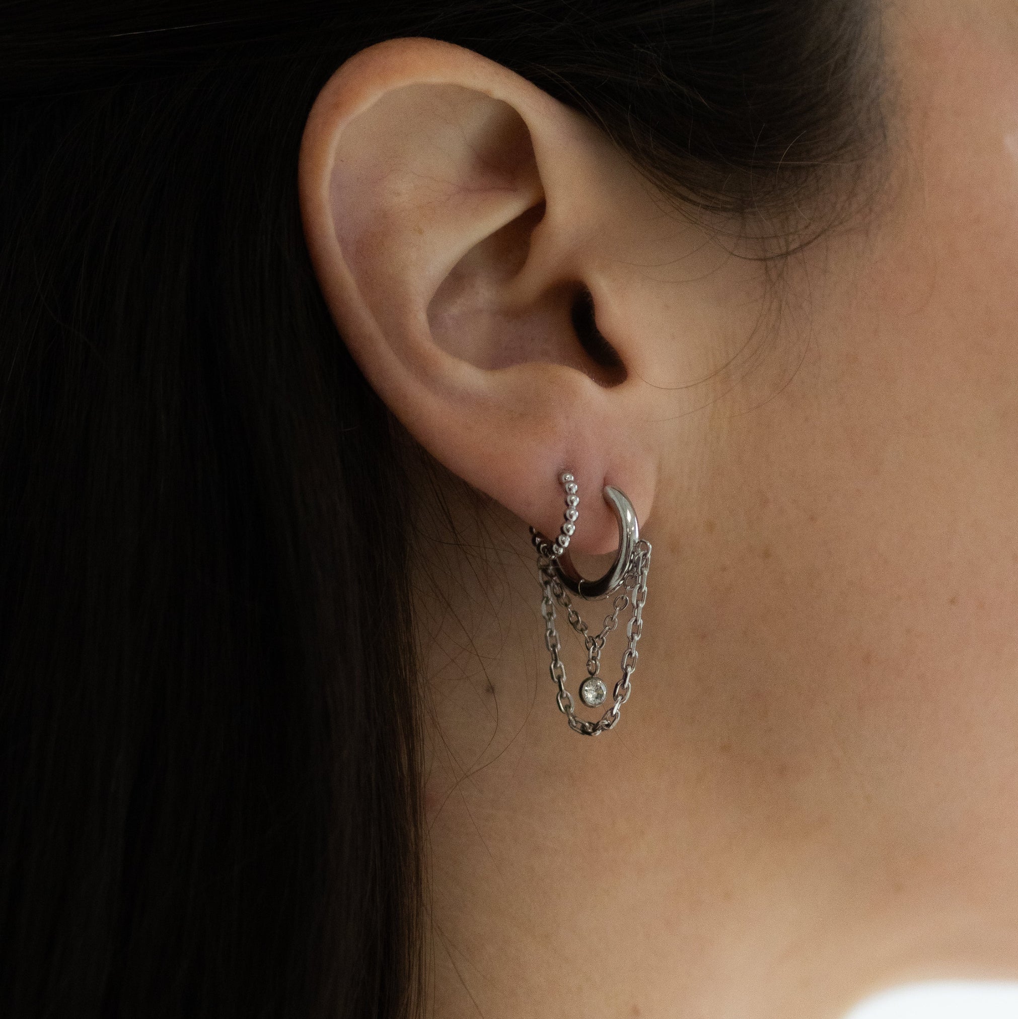 A close-up of a person's ear wearing a silver huggie hoop earring with dangling chain and pearl details.