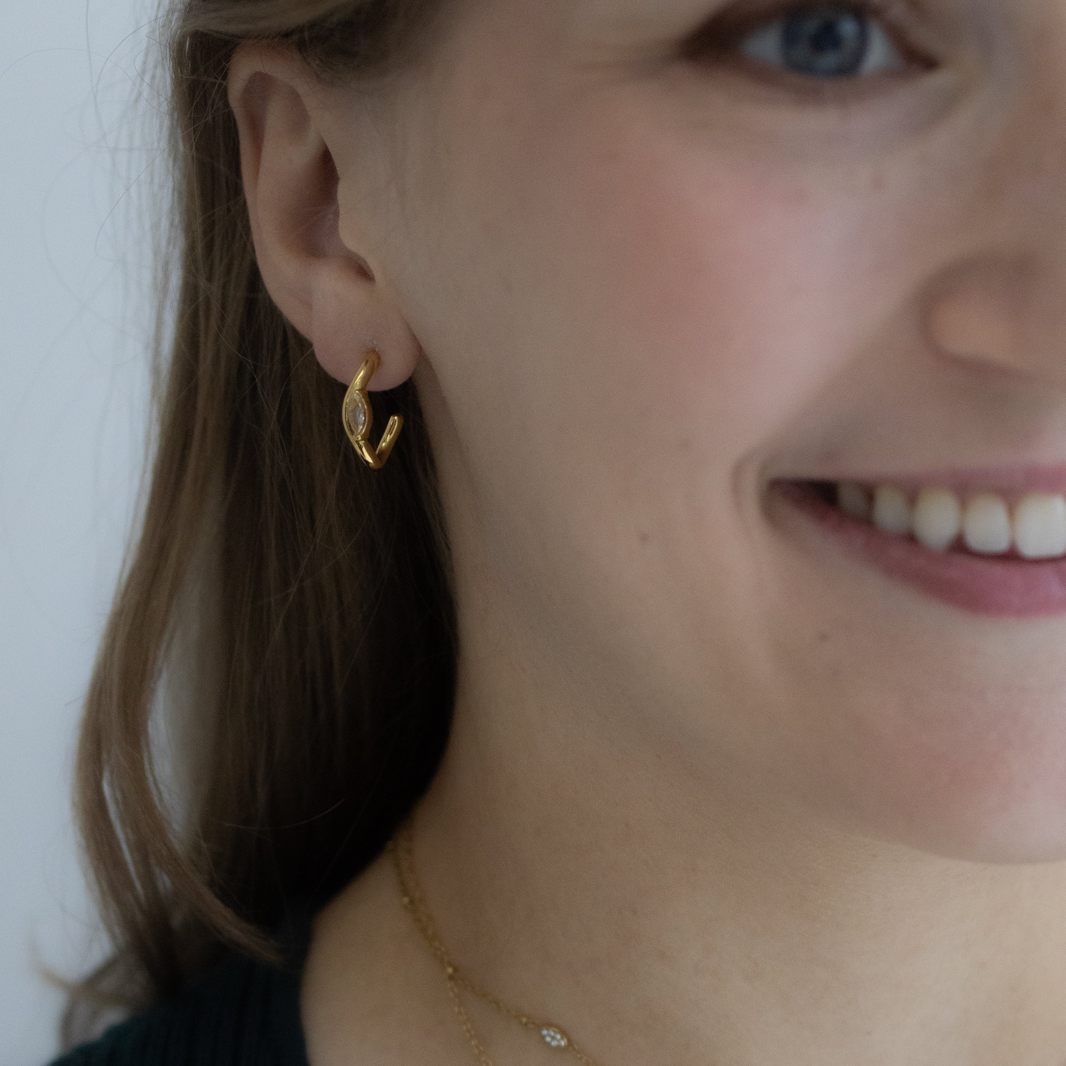 A close-up lifestyle shot of a woman wearing gold jewelry, featuring a small gold crescent moon earring and layered gold necklaces, including one with a small dangling pearl-like charm.