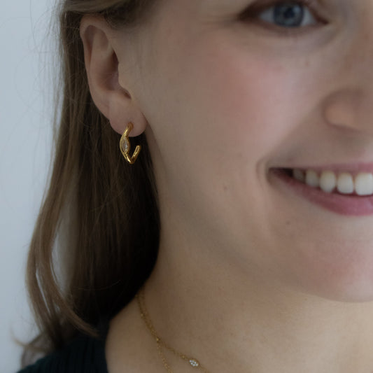 A close-up lifestyle shot of a woman wearing gold jewelry, featuring a small gold crescent moon earring and layered gold necklaces, including one with a small dangling pearl-like charm.