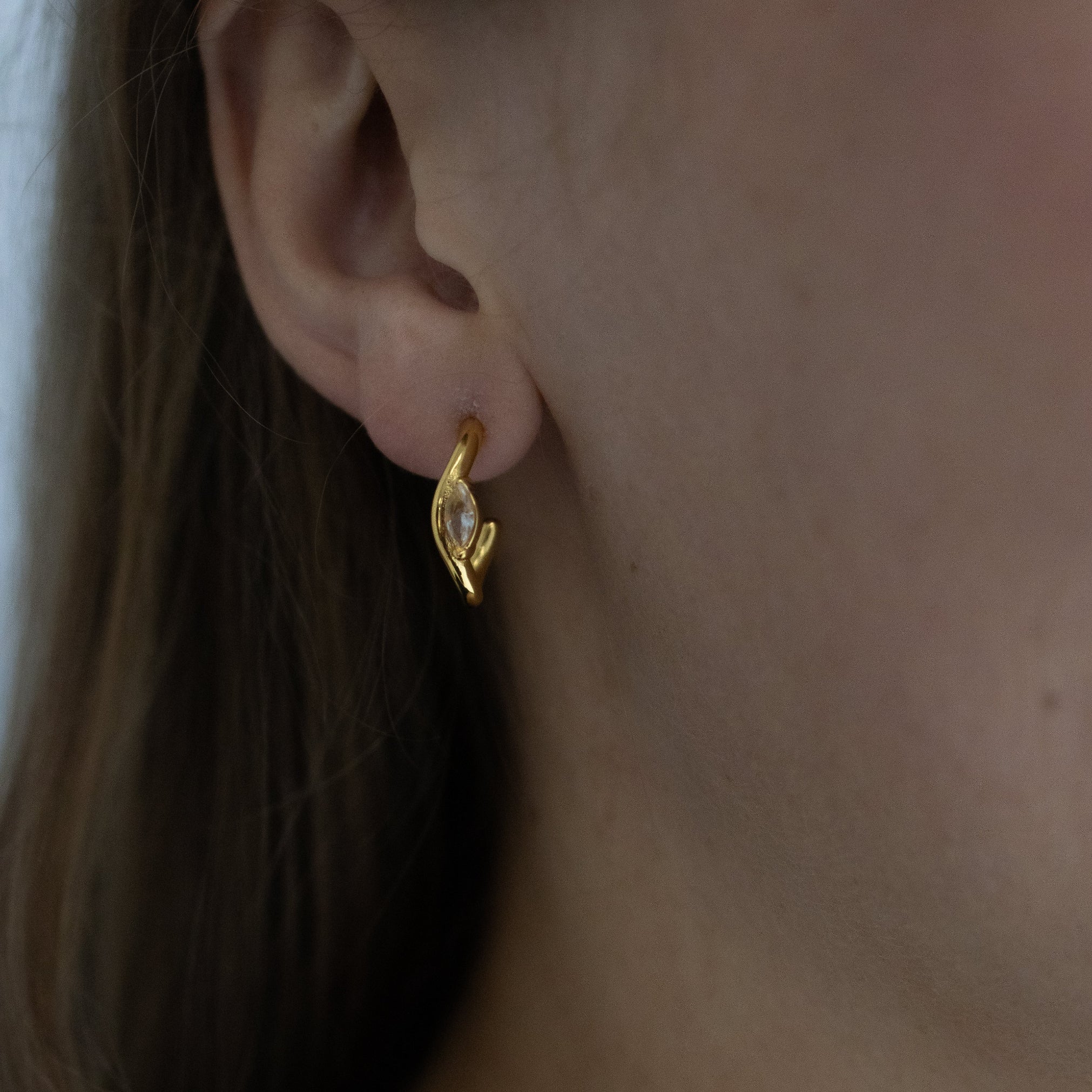 A close-up of a person's ear wearing a small gold huggie hoop earring with a clear faceted gemstone, along with a delicate gold chain necklace.