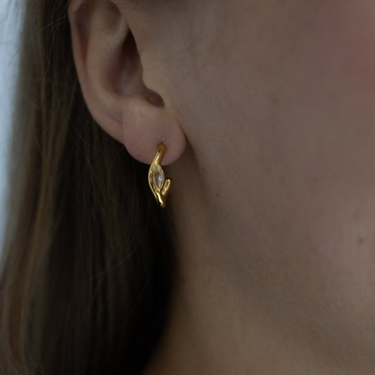 A close-up of a person's ear wearing a small gold huggie hoop earring with a clear faceted gemstone, along with a delicate gold chain necklace.